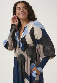 Long-sleeve dress with a mix of large blue, black, and beige abstract patterns; features smocked sleeves and a deep V-neckline.