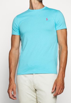 Mężczyzna noszący jasnoniebieski t-shirt z krótkim rękawem z małym różowym logo na lewej piersi oraz jasne spodnie.