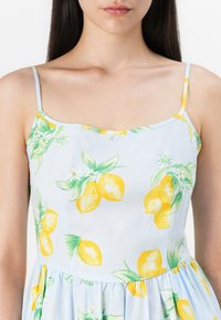 Blå ermeløs kjole med sitronmønster, har rund halslinning og rynket midje. Stoffet er glatt, lett og blomstermønstret.