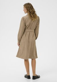 Brown corduroy dress with long sleeves and a belt at the waist. Mid-length, featuring vertical ribbed texture and a fitted silhouette.