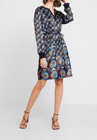 La robe noire présente un tourbillon coloré et un motif géométrique, des manches longues, un décolleté en V et une ceinture ajustable à la taille. Portée avec des talons à lanières.