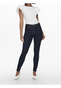 Mujer vestida con una blusa blanca de mangas abullonadas, jeans oscuros ajustados y unos zapatos de tacón blanco de punta, de pie contra un fondo sencillo.