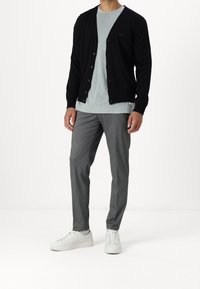 Černý kardigan s knoflíky, nošený přes světle modré tričko. Šedé kalhoty a bílé tenisky dokončují celý outfit. Jednoduchý, moderní styl.