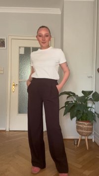 Άσπρη κοντομάνικη μπλούζα και σκούρα καφέ παντελόνι με φαρδιά πόδια και μπροστινές τσέπες. Απλός σχεδιασμός; το μοντέλο στέκεται σε έναν καλά φωτισμένο εσωτερικό χώρο.