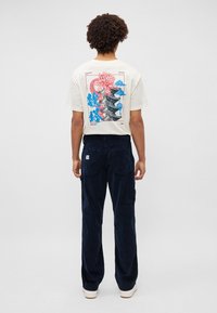 Tricou crem din bumbac cu un imprimeu grafic colorat pe spate. Pantaloni cargo din catifea albastră închisă, cu buzunare. Adidași albi.