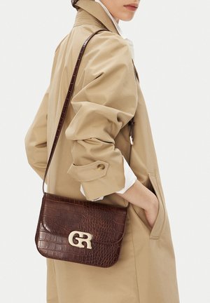 Žena v béžovém kabátě s vyhrnutými rukávy a s tmavě hnědou kabelkou s krokodýlím vzorem přes rameno, která má zlatou sponu "GR".