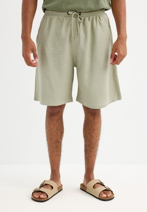 Hombre que lleva pantalones cortos de cordón verdes claros y sandalias beige, de pie sobre un suelo blanco contra un fondo sencillo.