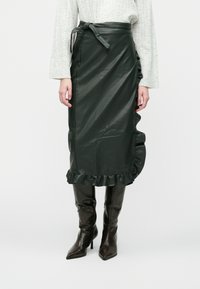 Dark green leather midi skirt with a ruffled hem, belted waist, and smooth texture. Paired with fitted, shiny brown knee-high boots.