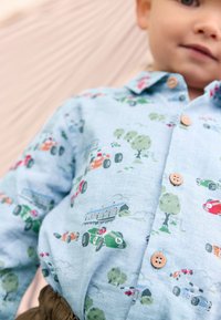 Camicia azzurra chiaro con bottoni che presenta un motivo di automobili e natura. Il materiale mostra una trama a spina di pesce. Include bottoni in legno e illustrazioni vivaci.