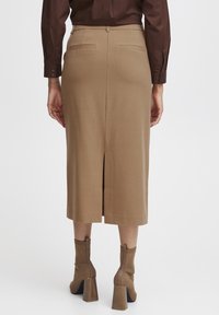 Knie-lange tan kleurige pencilrok met een split aan de achterkant, gecombineerd met bruin enkellange laarzen met hakken en een bruin shirt met lange mouwen.