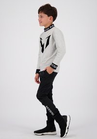 Sweatshirt gris avec logo "M" noir, manches noires avec texte "MESSI" blanc. Pantalons de sport noirs ; chaussures noires montantes avec semelles blanches.