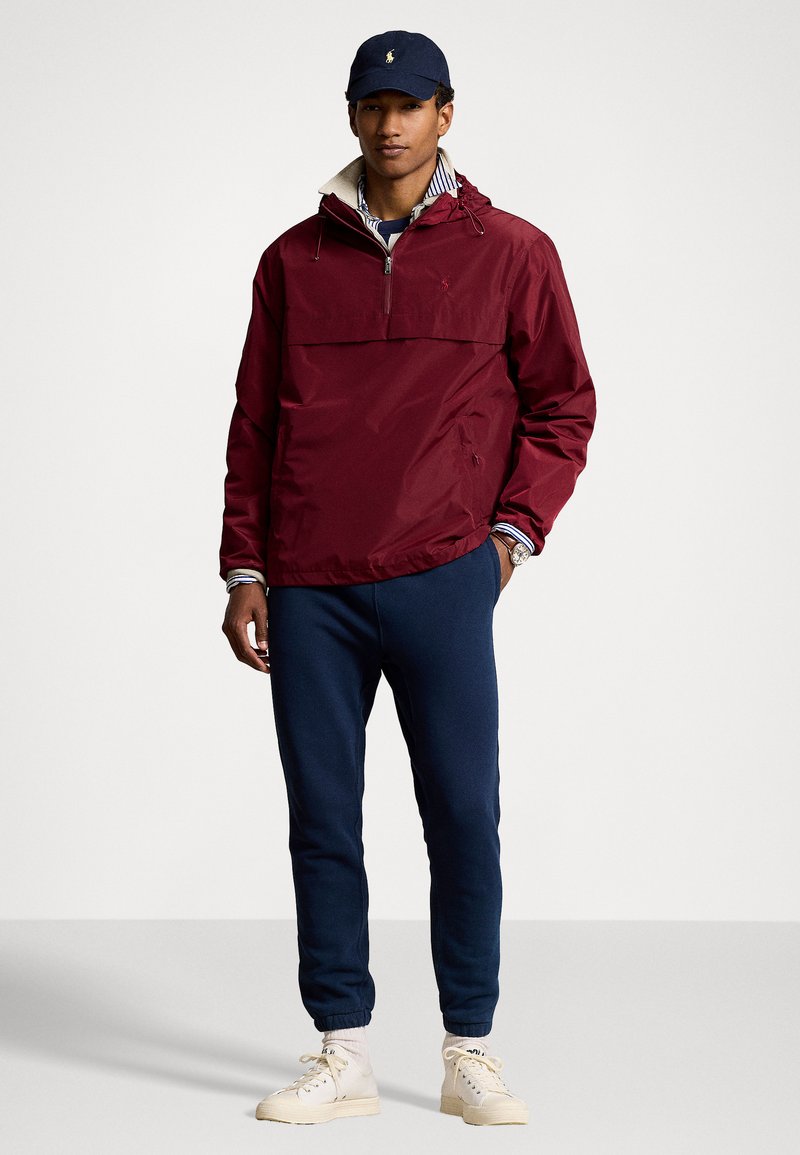 Bordeauxfarbener Pullover mit halbem Reißverschluss, mit Kordelzugkapuze; marineblaue Jogginghose; cremefarbene Sneaker; mit einem gestreiften Hemd darunter kombiniert.