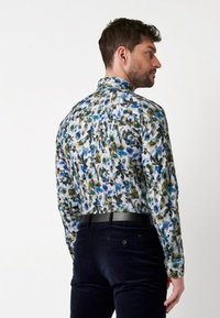 Bloemmotief shirt met blauwe bloemen en groene bladeren op een lichte achtergrond, met een kraag en lange mouwen, gecombineerd met donkere broek.
