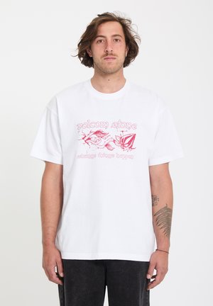 Homme aux cheveux mi-longs portant un T-shirt blanc avec un graphisme rouge et le texte « Volcom Stone » ainsi qu'un pantalon noir, debout devant un fond uni.