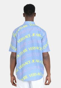 Camicia a maniche corte in tessuto blu chiaro con grandi stampe di testo giallo e rosa, tra cui "VERSACE" e "JEANS COUTURE."