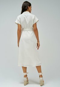 Vestido midi blanco con mangas cortas, cintura con cinturón y textura suave. Llevado con sandalias de cuña beige que tienen cintas en los tobillos.