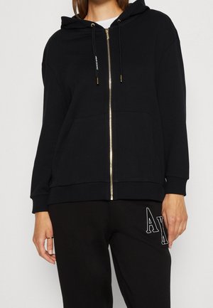 Osoba ubrana w czarną bluzę z kapturem zapinaną na zamek błyskawiczny z złotym zamkiem i sznurkami, w połączeniu z czarnymi spodniami z białymi literami "AX".