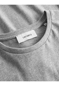 T-shirt cinza de algodão com gola redonda, apresentando uma textura lisa e uma etiqueta branca que diz "LES DEUX" cosida na gola.