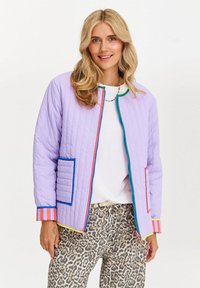 Blondynka uśmiechająca się, ubrana w jasnofioletową pikowaną kurtkę z kolorowymi wykończeniami, białą bluzkę i spodnie w leoparda na jednolitym tle.