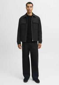 Man staat tegen een effen achtergrond in een zwart jack, een zwart kabelgebreid vest, wijde zwarte broek en zwarte schoenen.
