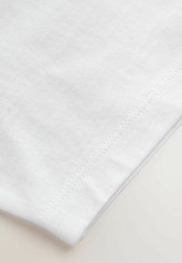 Weißer Baumwollstoff mit glatter Textur, der einen gestickten Saum aufweist und subtile Variationen in der Oberfläche zeigt. Der Rand ist sauber verarbeitet.