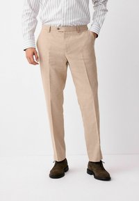 Pantalones de lino beige con corte recto, con pliegues frontales y cierre de un solo botón. Combinados con zapatos marrón oscuro.