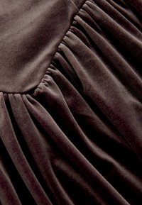 Temno žametna tkanina z gladko teksturo, s nabranimi gubami, ki ustvarjajo vizualno globino in mehkobo. Bogata, temna barva poudarja oblikovne elemente.