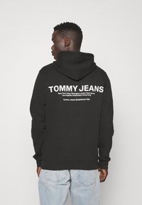 Felpa nera con un grande logo bianco "TOMMY JEANS" sul retro, dotata di tasca a marsupio e cappuccio con stringa. Sono indossati jeans azzurri chiari.