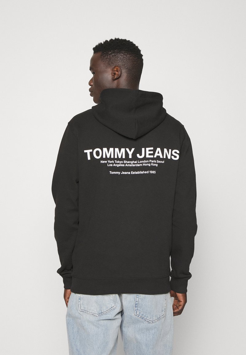 Felpa nera con un grande logo bianco "TOMMY JEANS" sul retro, dotata di tasca a marsupio e cappuccio con stringa. Sono indossati jeans azzurri chiari.