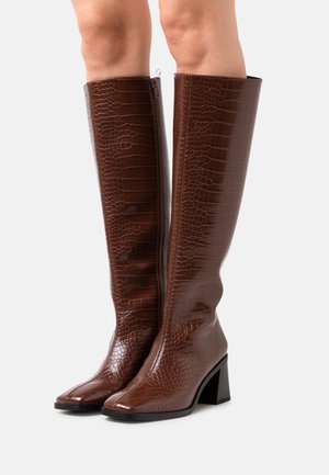 Kniehoge bruine nep-krokodillenleren laarzen met blokhakken, gedragen op blote benen tegen een effen achtergrond.