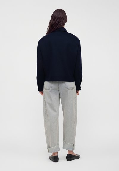 Personne aux longs cheveux porte une veste noire, un jean gris clair à revers et des chaussures plates noires, debout face à un fond blanc uni.
