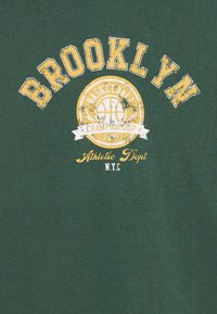 T-shirt en coton vert avec "BROOKLYN" en lettres jaunes en gras, et un graphique de basketball circulaire avec "CHAMPIONNAT DE BASKETBALL 1999" en dessous.