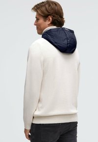 Hellcremefarbener Pullover aus strukturiertem Stoff mit gerippten Bündchen und einer marineblauen gesteppte Kapuze. Von hinten gezeigt, getragen zu dunkelgrauen Jeans.