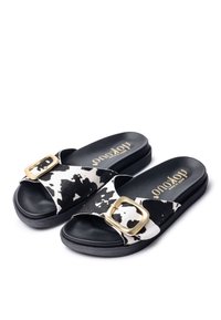 Black and white cow print slides with a wide strap featuring a gold-tone buckle. Textured footbed and sturdy black sole.