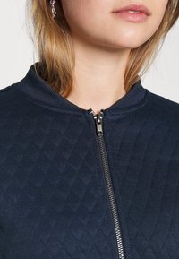 Granatowa pikowana kurtka na zamek z prążkowanym kołnierzem i srebrnym suwakiem. Widoczny fakturowany wzór na tkaninie dodaje głębi. Prezentacja z bliska.