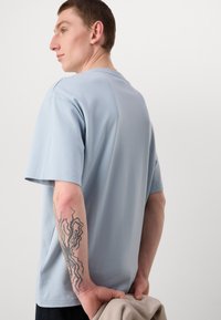Light blue short-sleeve t-shirt made of soft fabric, featuring a round neckline. A tattoo is visible on the model's forearm.