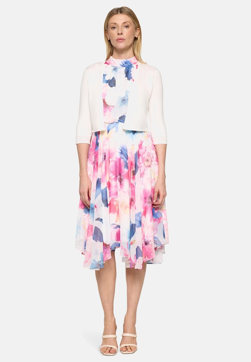 Femme debout portant un cardigan blanc court sur une robe fleurie aux motifs roses, bleus et blancs, atteignant les genoux, avec des sandales blanches à brides.