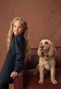 Vestito camicia con stampa maculata nera e maniche lunghe; ragazza con capelli biondi ondulati accanto a un cane soffice marrone chiaro su un divano in pelle marrone.