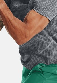 Camicia a maniche corte grigia con motivo a quadri, abbinata a pantaloni corti verdi. Il tessuto sembra leggero e con una texture.