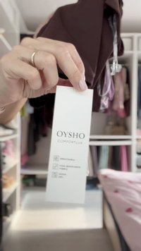 Vit etikett som visar "OYSHO COMFORTLUX." Med symboler för andningsbara, vattentåliga och snabbtorkande tyger. Bakgrunden inkluderar kläder.