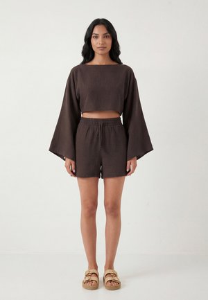 Vrouw staat, gekleed in een donkerbruine losse crop top met lange mouwen en bijpassende korte broek, met beige sandalen tegen een effen witte achtergrond.