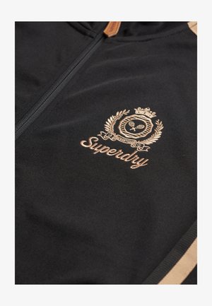 Sort zip-up jakke lavet af glat stof med et guld broderet logo og detaljer, med kontrasterende tan striber på ærmerne.