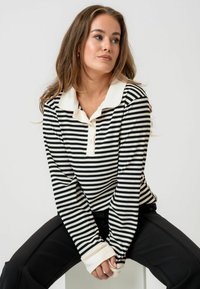 Crno-bijela prugasta majica s dugim rukavima i kremastim ovratnikom te dugmadi. Mekana rebrasta tekstura s produženim manžetama.