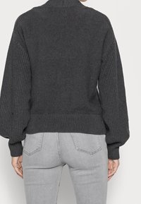 Žena v tmavě šedém hrubém pleteném svetru s nafouklými rukávy a světle šedých přiléhavých džínách, zobrazena zezadu.