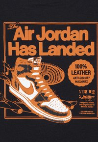 Juodas marškinėlis su oranžiniu grafiniu piešiniu aukštakulnio sportinio bato, pažymėtu "Air Jordan Has Landed", su tekstūruotu raštu ir drąsiais tekstais.
