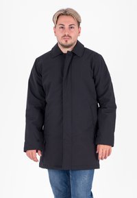 Cappotto nero realizzato in tessuto liscio, dotato di collare, chiusura con bottoni e tasche laterali. Indossato sopra jeans blu, il modello è in posa su uno sfondo bianco.