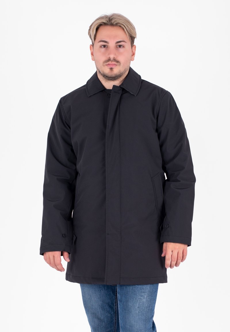 Cappotto nero realizzato in tessuto liscio, dotato di collare, chiusura con bottoni e tasche laterali. Indossato sopra jeans blu, il modello è in posa su uno sfondo bianco.