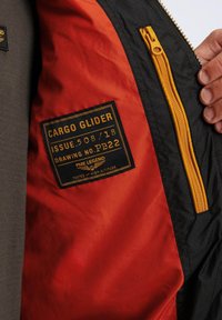 Zwarte jas interieur met een oranje voering, gele rits en een zwart label met gouden tekst waarop het model en het versienummer staan.