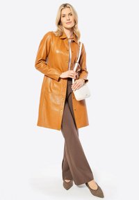 Tan leren jas met een kraagontwerp, knopen aan de voorkant, twee zakken. Gepaard met een bruine broek en een witte, gewatteerde schoudertas.