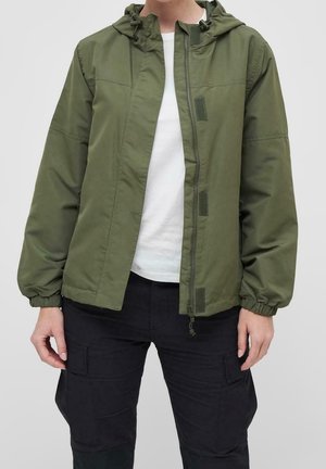Personne portant une veste à capuche vert olive sur une chemise blanche et un pantalon cargo noir, vue des épaules jusqu’aux hanches.